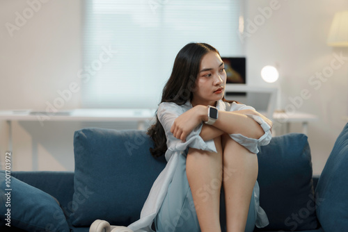 Fotografie Young asian woman is sitting on a sofa at home, hugging her knees and looking aw
