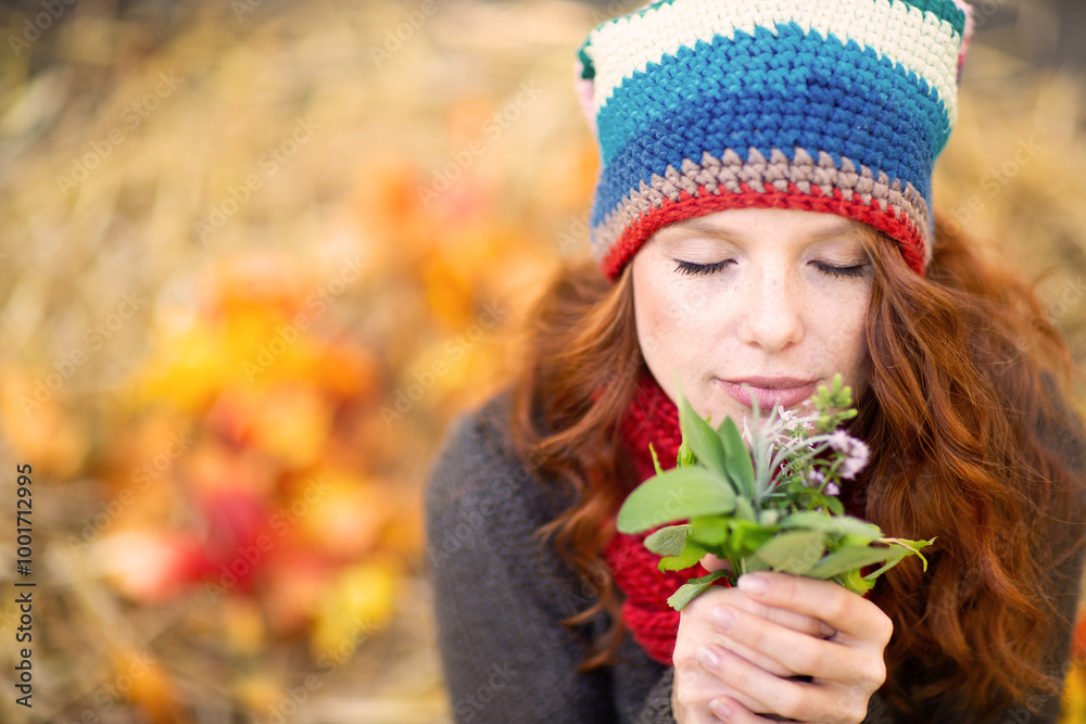 Fototapeta premium Serene woman with red hair, wearing a colorful knit hat and gray sweater, holds a small bouquet of flowers close to her face, eyes closed, enjoying the fragrance, surrounded by warm autumn colors.