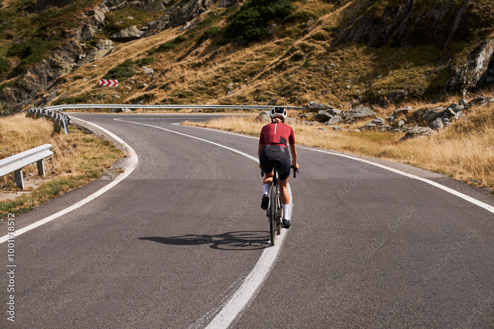 Fototapeta premium Woman cyclist riding up a winding mountain road, showcasing the beauty of the natural environment. Sport motivation.Female cyclist wearing a cycling kit and helmet. Carpathian Mountains,Transfăgărășan
