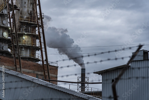 A large industrial facility releases smoke into the gray sky, showcasing the complexity of urban life amid towering structures and barbed wire photo