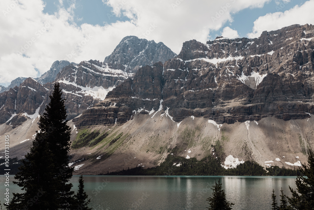 Obraz premium view of a rocky cliff with snow patches next to a calm lake with clouds in the sky in summer with trees in the foreground