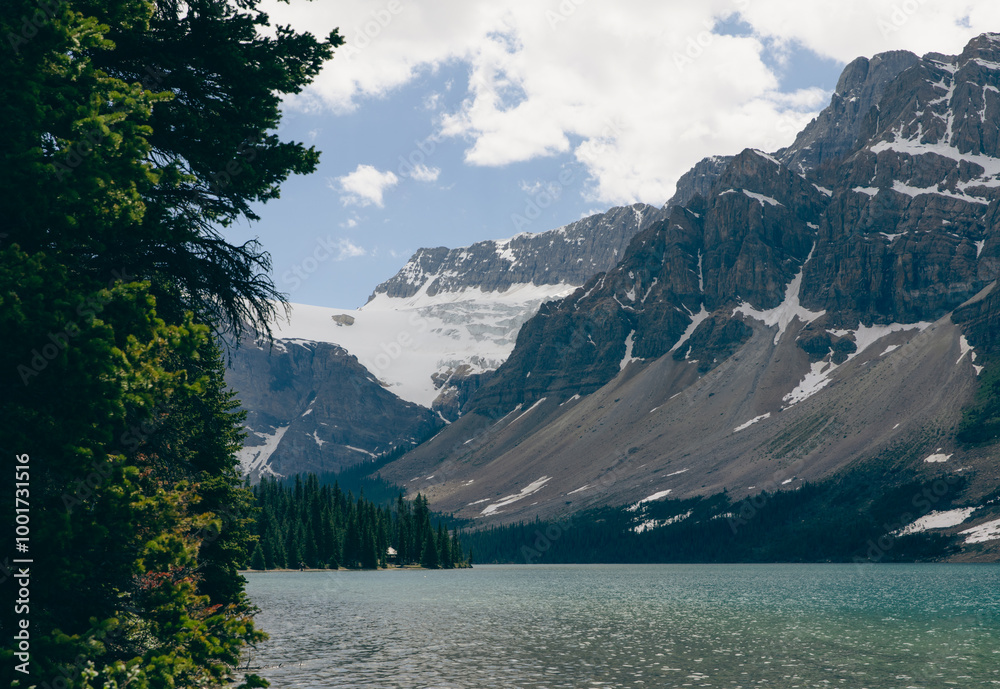 view of a rocky cliff with snow patches next to a calm lake with bright clouds in the sky in summer view of a rocky cliff with snow patches next to a calm lake with bright clouds in the sky in summer