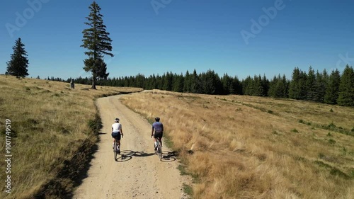 Two cyclists ride on a dirt road through a grassy field with a forest in the background on a sunny day. .Cycling on gravel roads or off-road.Active adventures and the concept of travel by bike. 