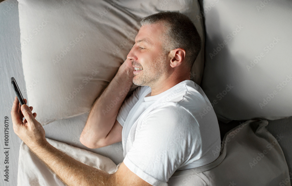 Smiling middle aged man using smartphone while lying in bed, texting and browsing internet resting on pillow in bedroom interior, top view shot Smiling middle aged man using smartphone while lying in bed, texting and browsing internet resting on pillow in bedroom interior, top view shot