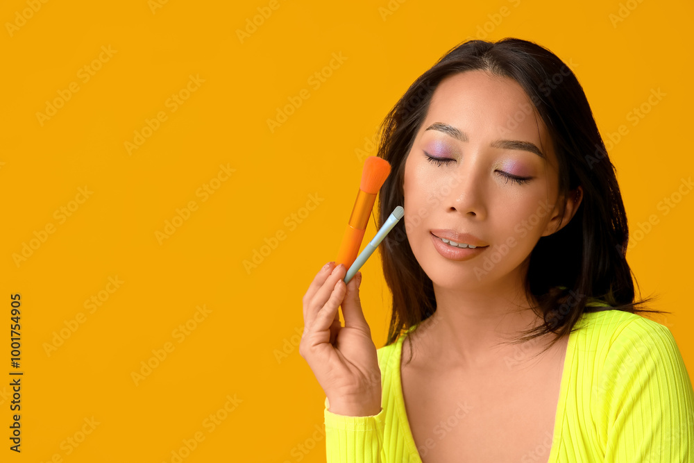 Young Asian woman with makeup brushes on yellow background Young Asian woman with makeup brushes on yellow background