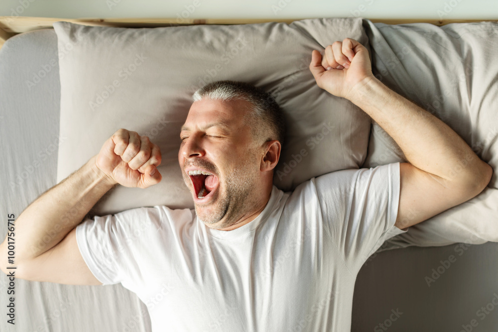 Good morning. Rested middle aged man waking up, yawning and stretching hands while lying in bed, top view shot Good morning. Rested middle aged man waking up, yawning and stretching hands while lying in bed, top view shot