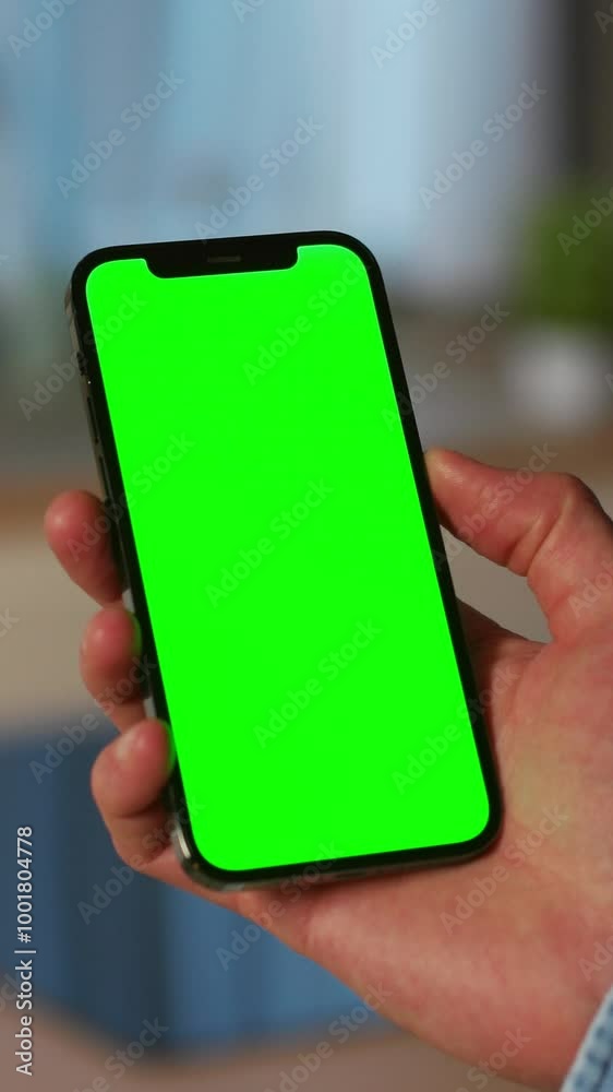 Shot of man's hand wearing jeans jacket holding smartphone with green screen in vertical position isolated on blurred background of kitchen. Unrecognizable man reading dinner ideas on Internet Shot of man's hand wearing jeans jacket holding smartphone with green screen in vertical position isolated on blurred background of kitchen. Unrecognizable man reading dinner ideas on Internet