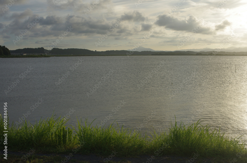 Fototapeta premium 汽水湖の涸沼に夕陽が静かに沈んでいく。広大な涸沼ラムサール湿地の一日の終わり。雄大な景色。刻々と変化する湖面の輝き。自然が織りなす光景に心を奪われ、茫然と佇んでいる私。一瞬、不安な気持ちに襲われた。温暖化が進行している地球、大丈夫か地球。地球が熱くなる。母なる地球が、今、悲鳴を上げている。私たちは何をなすべきか、できることは何か。
