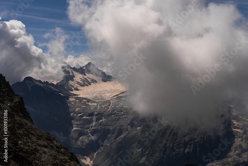 Dramatic mountain scene with cloud-covered peaks, snow patches, and rugged glacial terrain. The interplay of light and shadow adds depth to this breathtaking alpine landscape