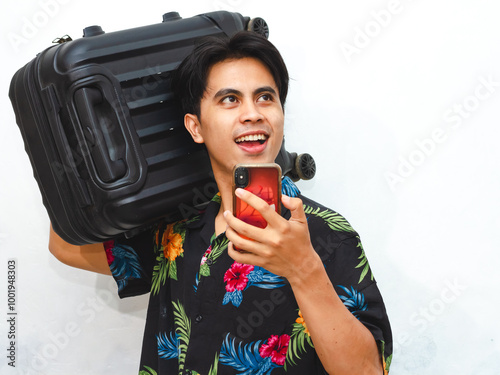 Portrait of a happy young Asian man wearing casual summer clothes, holding a mobile phone and suitcase, looking aside thoughtfully, representing a traveler ready for an adventure. photo