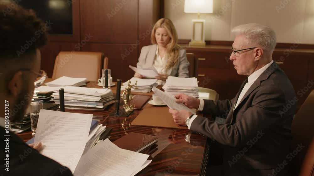 Senior grey-haired law firm leader giving documents to two colleagues sitting next to him at wooden table at meeting in boardroom Senior grey-haired law firm leader giving documents to two colleagues sitting next to him at wooden table at meeting in boardroom