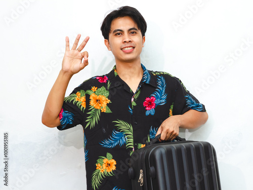 Portrait of a young Asian man in summer casual clothes, holding a suitcase, smiling happily, giving a thumbs-up and OK sign, ready for travel or vacation. Isolated on a white background. photo