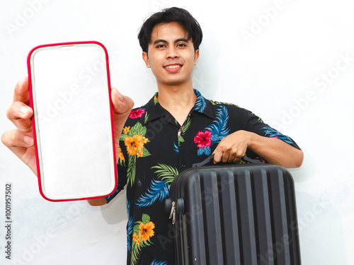 Young Asian man in summer casual wear joyfully shows a blank mobile phone screen while carrying a suitcase, embodying the spirit of travel and adventure. photo