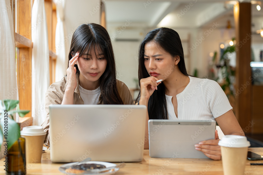 Two serious and stressed Asian female friends are working remotely together from a coffee shop. Two serious and stressed Asian female friends are working remotely together from a coffee shop.