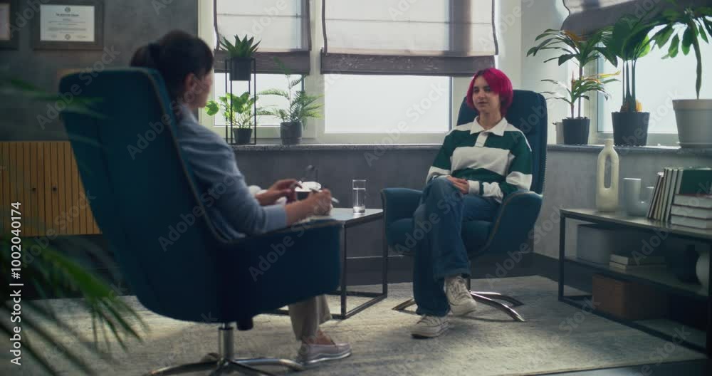 In psychotherapy room, teenage girl talks to her therapist, shares positive emotions during counseling session. Back view of female psychologist sitting in chair, providing therapy for to teenager.