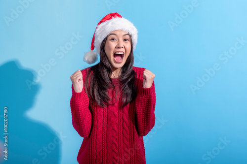 An Asian woman wearing a red knit sweater and a Santa hat celebrates with a joyful expression. Her hands are clenched into fists and raised in excitement. The background is light blue.