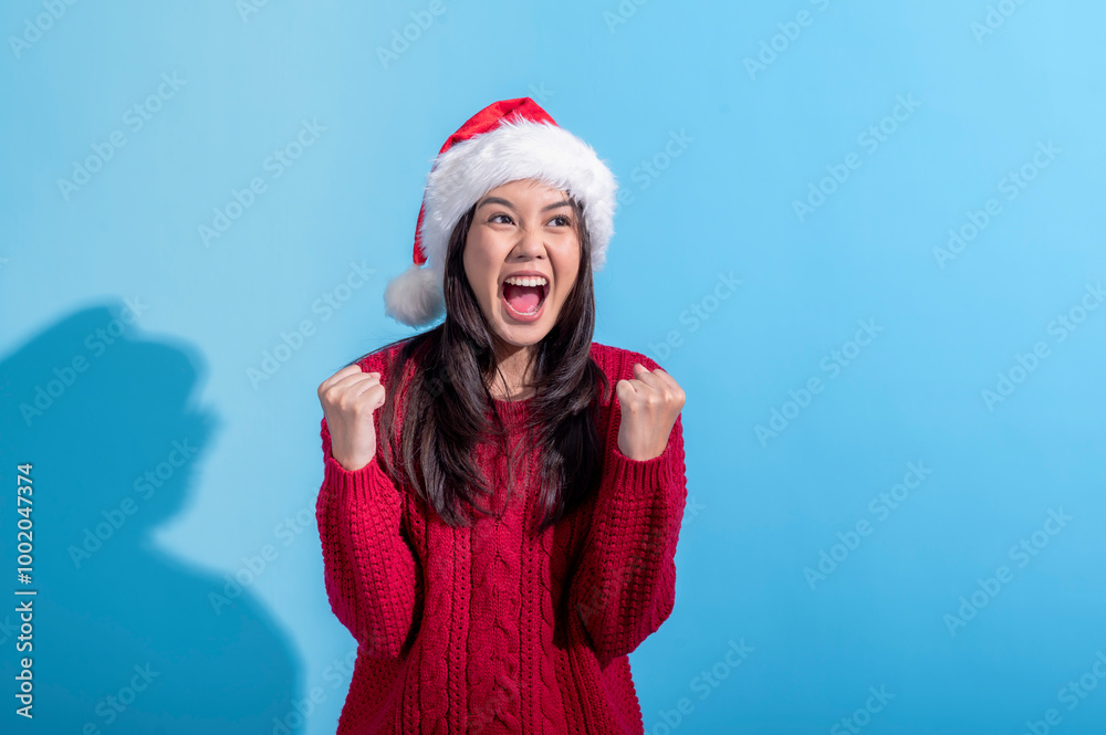 An Asian woman wearing a red knit sweater and a Santa hat celebrates with a joyful expression. Her hands are clenched into fists and raised in excitement. The background is light blue. An Asian woman wearing a red knit sweater and a Santa hat celebrates with a joyful expression. Her hands are clenched into fists and raised in excitement. The background is light blue.