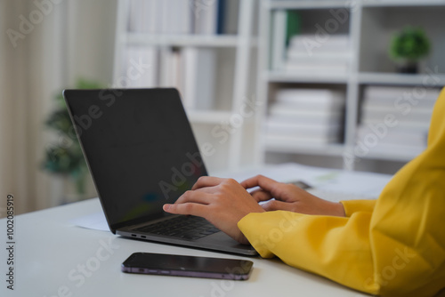 Focused on the Task: A close-up shot of a woman's hands typing on a laptop, showcasing a modern workplace setting with a bookshelf in the background, symbolizing focus, productivity.