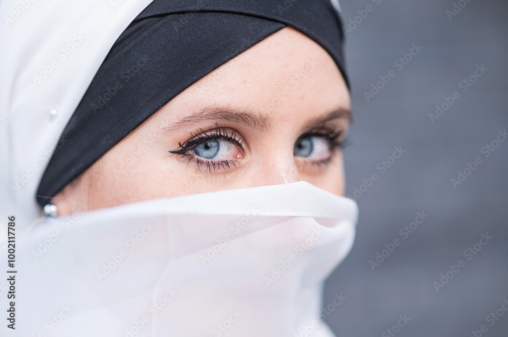 Portrait of a young blue-eyed woman in a hijab against a gray brick wall. Muslim woman with white scarf covering her face. Portrait of a young blue-eyed woman in a hijab against a gray brick wall. Muslim woman with white scarf covering her face.