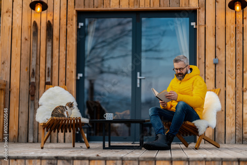 Mature man sitting on terrace reading book, enjoying cup of coffee during cold winter day. Peaceful moment alone in cabin. photo