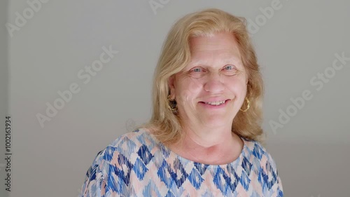 A blonde mature woman smiling warmly at the camera in a comfortable home setting, radiating positivity and warmth.