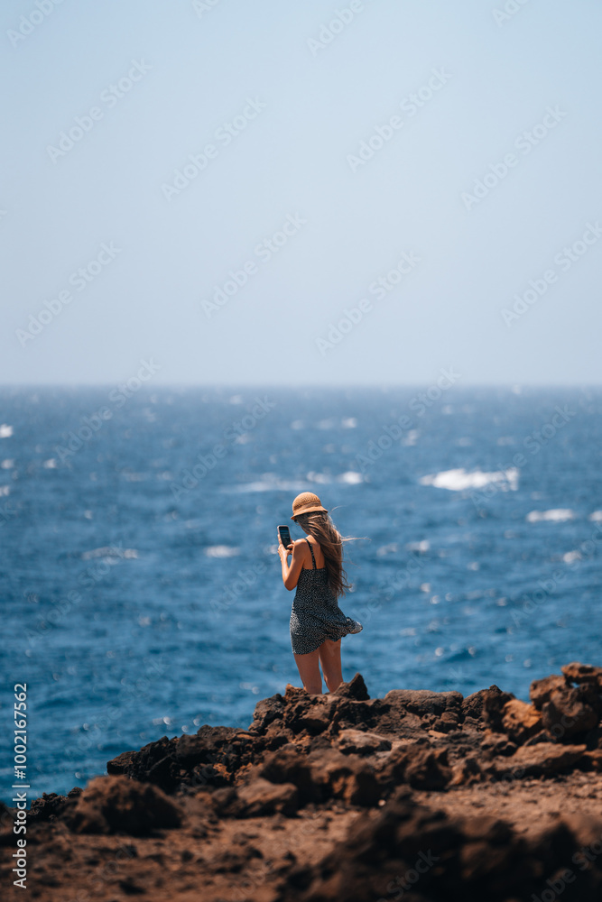 A woman stands on rocky coastline taking photos with her smartphone of the ocean
