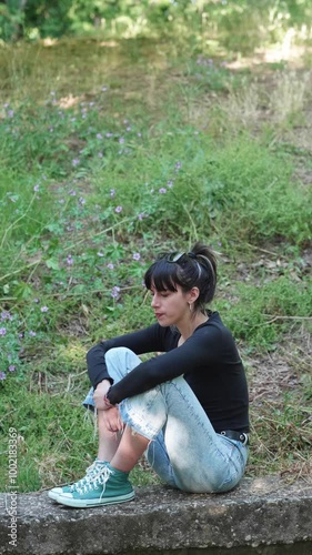 Vertical. A brunette woman with straight hair wearing a black shirt sits on a wall in the park, looking sad and despondent because she lost her job, capturing a moment of sorrow.