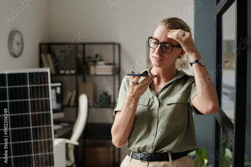 Young blond businesswoman in eyeglasses recording voice message on mobile phone against solar panel photo