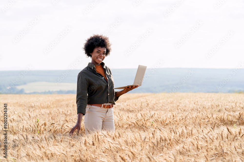 Smiling Afro agronomist with laptop amidst barley crops in field Smiling Afro agronomist with laptop amidst barley crops in field