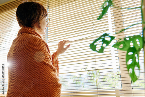 Mature woman wrapped in blanket looking through window blinders at home