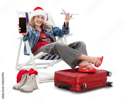 Christmas Holiday travel and vacation concept, tourist woman in travel attire wears a santa hat, on deck chair with trolley suitcases, showing smartphone and model airplane. Flight and travel booking.