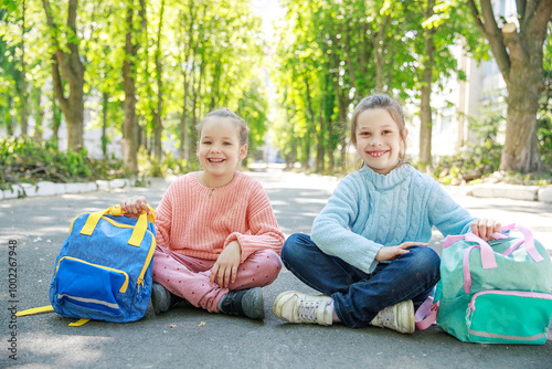 Two joyful girls sit cross-legged on a quiet street, basking in the sunlight. The vibrant greenery around them enhances their playful spirit, making for a delightful afternoon.