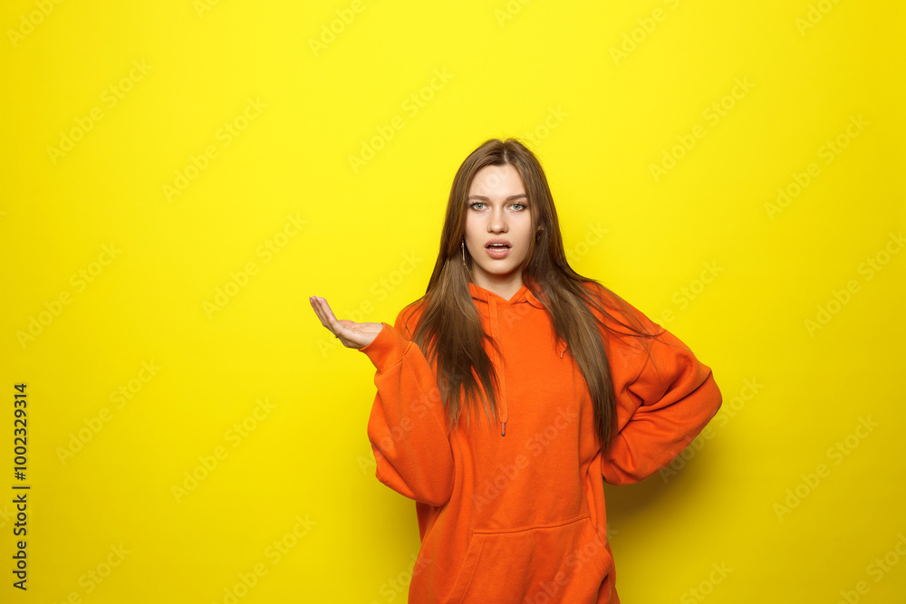 Young woman in orange hoodie on yellow background, surprised, pointing with hand. Young woman in orange hoodie on yellow background, surprised, pointing with hand.