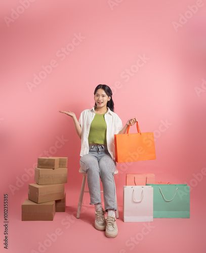 Portrait of Asian woman posing on pink background, next to shopping bags.