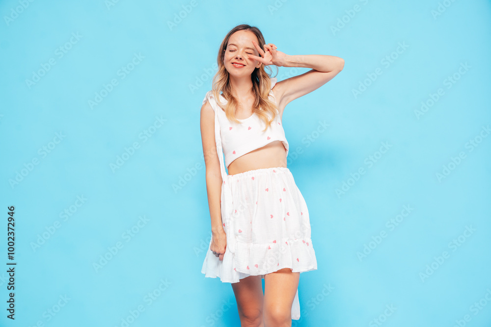 Young beautiful smiling blond female in trendy summer white dress clothes. Carefree woman posing near blue wall in studio. Positive model in. Cheerful and happy Young beautiful smiling blond female in trendy summer white dress clothes. Carefree woman posing near blue wall in studio. Positive model in. Cheerful and happy