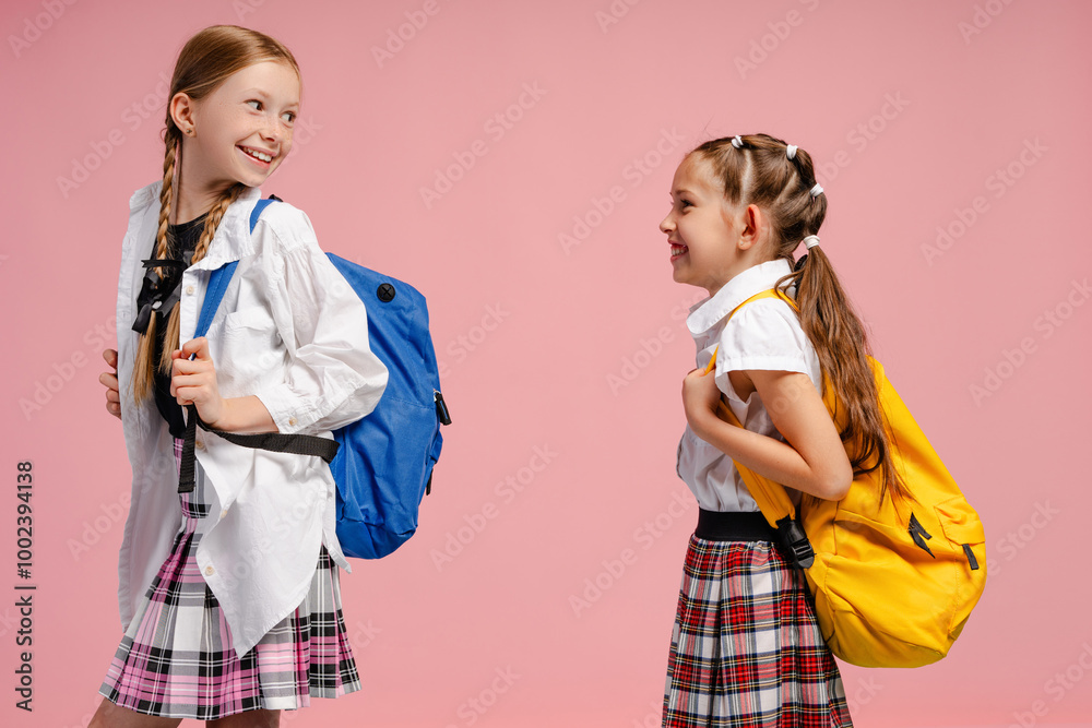 Two smiling schoolgirls wearing backpacks and talking on pink background Two smiling schoolgirls wearing backpacks and talking on pink background