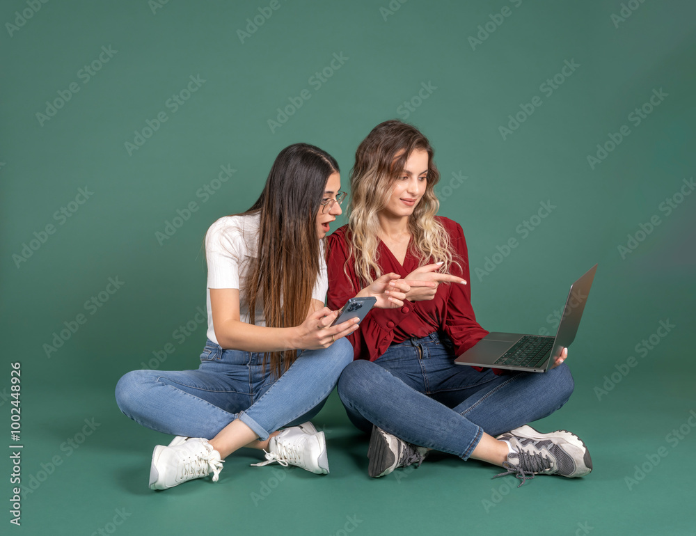 Full body length two young caucasian women girl friends sit floor over green background hold use pointing laptop screen with positive gesture. Speaking, meeting online working concept idea image. Full body length two young caucasian women girl friends sit floor over green background hold use pointing laptop screen with positive gesture. Speaking, meeting online working concept idea image.