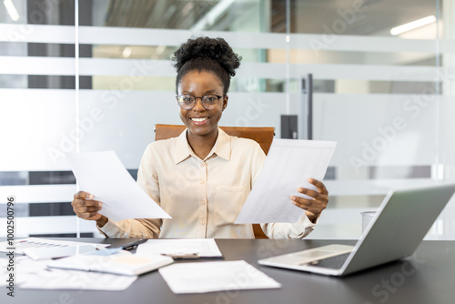 Confident businesswoman smiles while holding documents in office. She reviews her work at modern desk with laptop, showcasing professionalism and positivity.