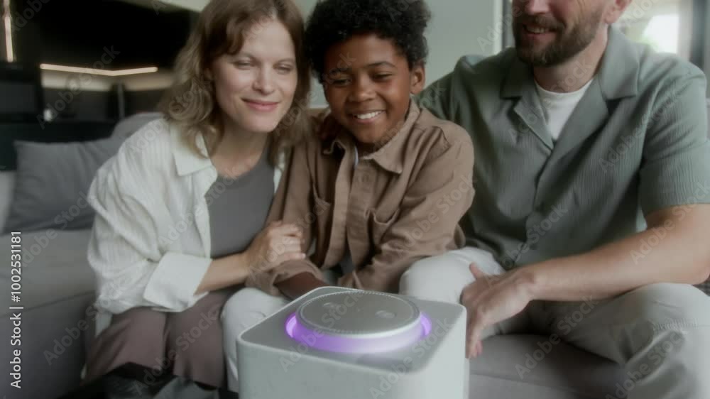 Medium shot of young black boy sitting on couch with foster Caucasian parents at new home, pressing button on interactive smart speaker, saying command, listening to response and laughing Medium shot of young black boy sitting on couch with foster Caucasian parents at new home, pressing button on interactive smart speaker, saying command, listening to response and laughing
