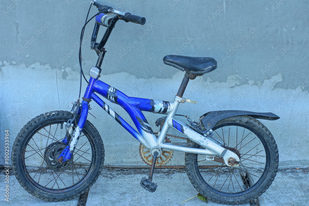 one children's iron modern blue individual sports small with two black wheels industrial with handlebars and soft saddle bicycle stands near gray concrete wall during daytime on the street one children's iron modern blue individual sports small with two black wheels industrial with handlebars and soft saddle bicycle stands near gray concrete wall during daytime on the street