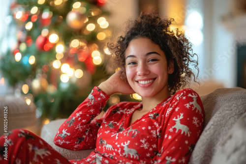 Young woman is smiling while relaxing on the sofa wearing a festive christmas pajamas in front of a decorated christmas tree