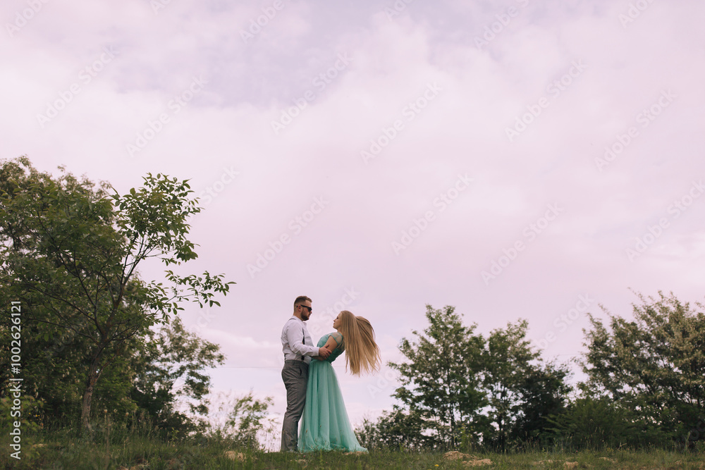 A couple is standing in a field with trees in the background. The woman is wearing a green dress and the man is wearing a white shirt. They are holding hands and smiling at the camera A couple is standing in a field with trees in the background. The woman is wearing a green dress and the man is wearing a white shirt. They are holding hands and smiling at the camera