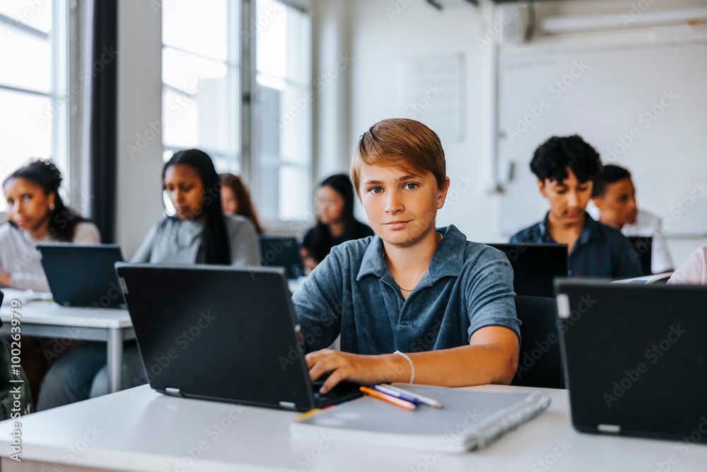 Portrait of teenage boy sitting with laptop near desk in classroom at school Portrait of teenage boy sitting with laptop near desk in classroom at school