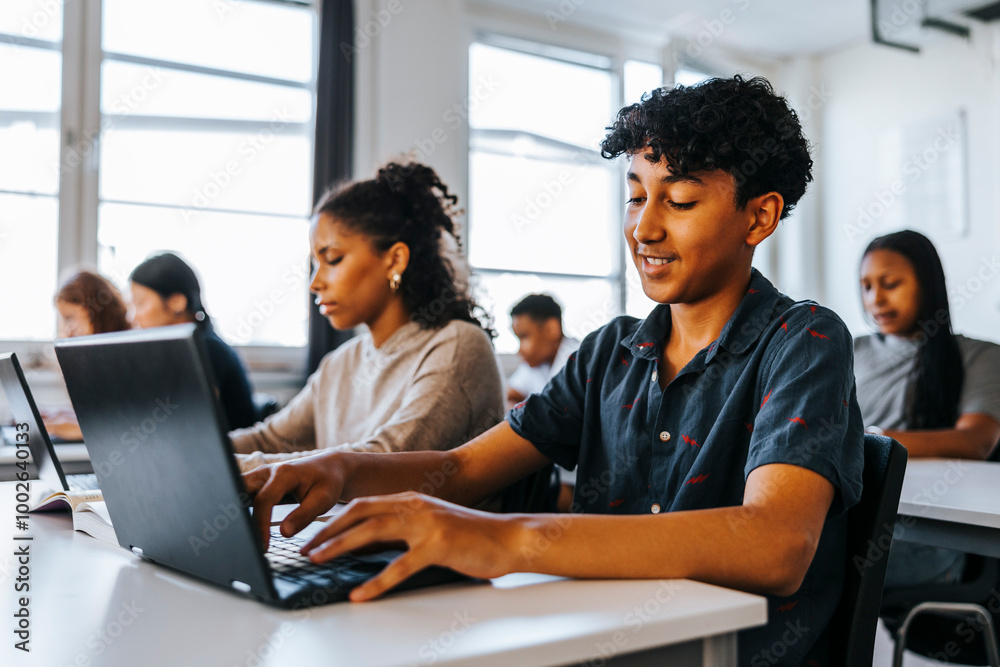 Smiling teenage boy using laptop while sitting with students in classroom at junior high school Smiling teenage boy using laptop while sitting with students in classroom at junior high school