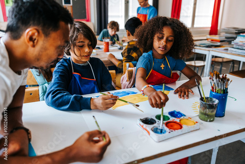 Art professor leaning on desk and teaching pupils painting while sitting in classroom at school