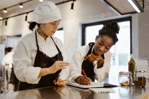 Female chef with writing while discussing with colleague standing in commercial kitchen photo