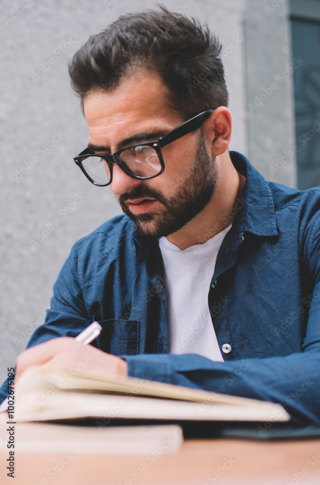 Concentrated male student in classic spectacles for vision correction sitting at street cafeteria and learning preparing to university exams, bearded man making memo notes in knowledge textbook Concentrated male student in classic spectacles for vision correction sitting at street cafeteria and learning preparing to university exams, bearded man making memo notes in knowledge textbook