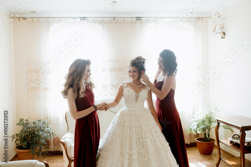 Three women are getting ready for a wedding. One of them is touching the bride's hair. The room is decorated with a potted plant and a vase photo