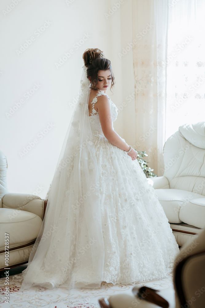 A woman in a white wedding dress stands in front of a white couch. She is wearing a veil and a necklace A woman in a white wedding dress stands in front of a white couch. She is wearing a veil and a necklace
