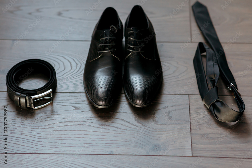 A pair of black shoes and a belt are on a wooden floor. The shoes are placed in front of the belt, and the belt is hanging on the floor. Concept of organization and preparation, as the shoes A pair of black shoes and a belt are on a wooden floor. The shoes are placed in front of the belt, and the belt is hanging on the floor. Concept of organization and preparation, as the shoes